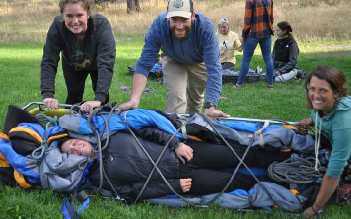 a student lies bundled in a stretcher while three others smile at the camera during a wilderness first aid training